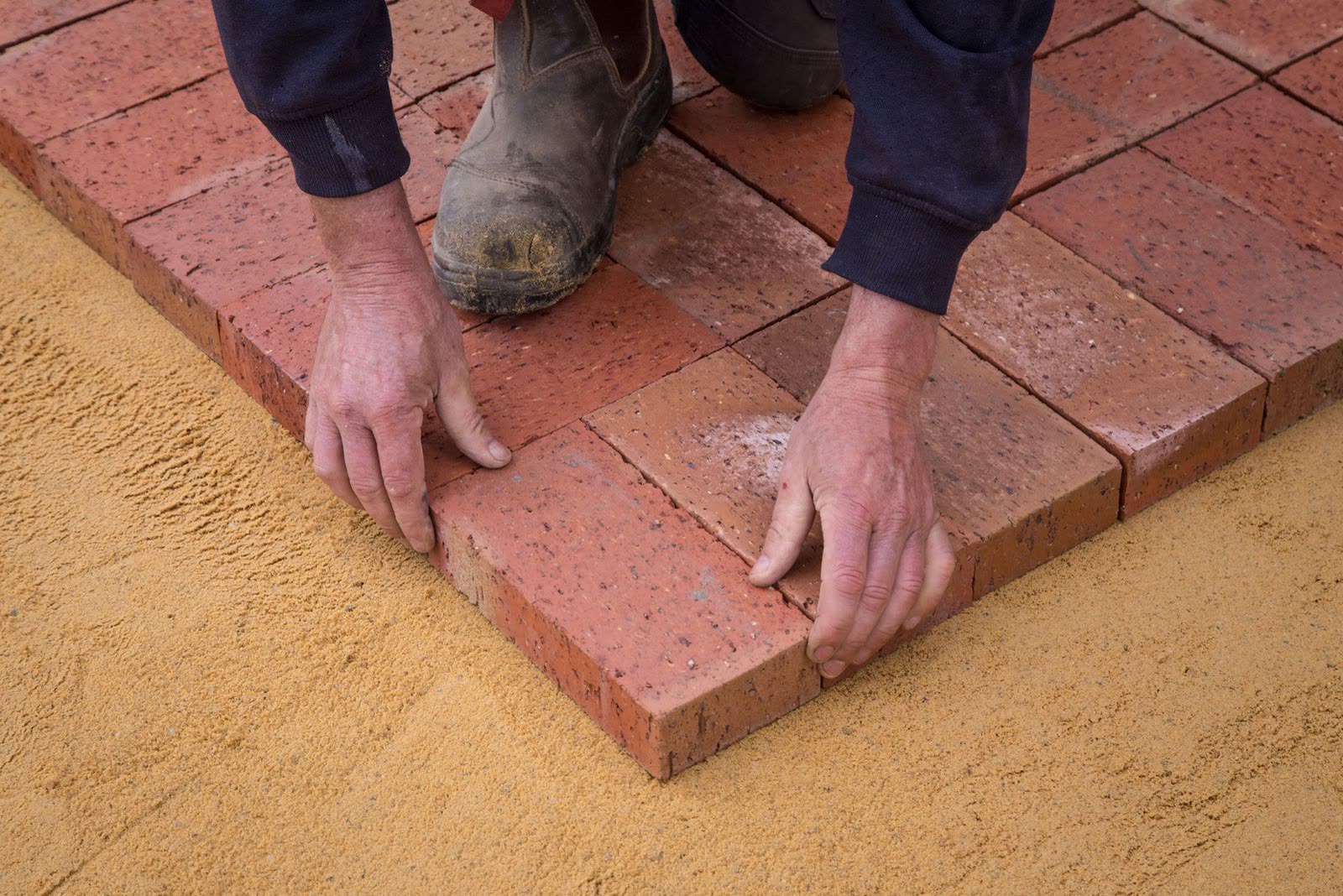 Hands setting red brick pavers into a leveled sand bed